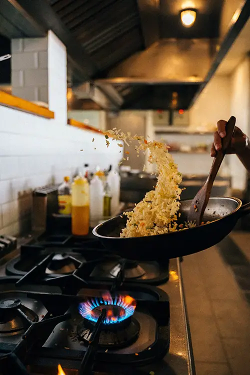 Un chef faisant sauter du riz dans une marmite dans une cuisine de restaurant, révélant des bouteilles de sauces et d'huile sur son comptoir.
